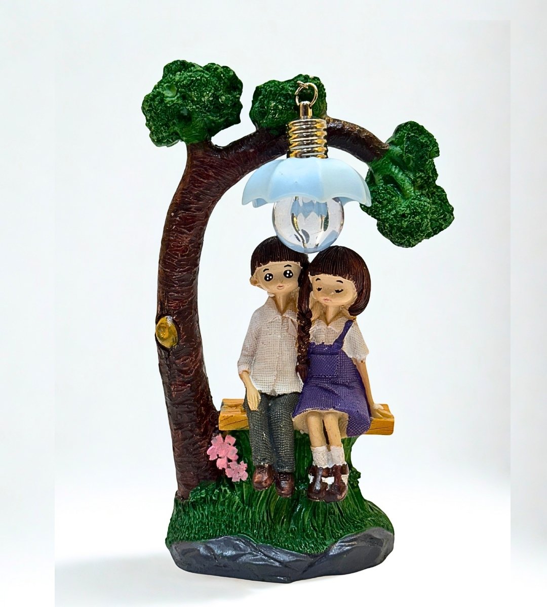 Cute figurine of a boy and girl sitting under a flower-shaped lamp on a bench beside a tree